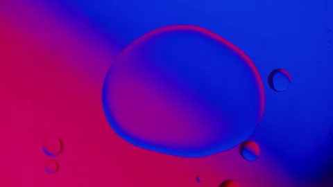 A large drop absorbs several smaller drops on a blue-red gradient background. 動画素材 303058570