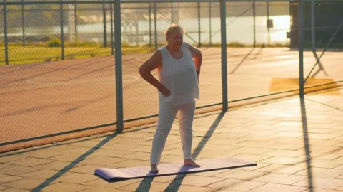 Large elderly lady doing hip rotation exercises on outdoor mat. fitness and Stock Footage 340993725