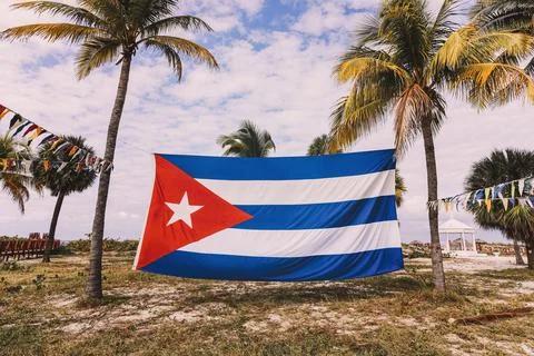 A large flag of Cuba is strung between palm trees, on the beach of the Caribb Stock Photos