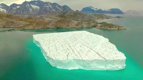 Large flat iceberg floating in arctic waters near rocky coastline under cloudy Stock Footage 343695687