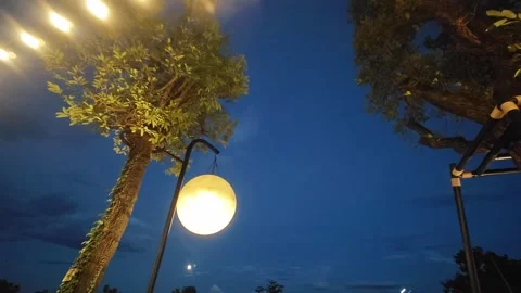 Large glowing moon replica hanging from a black pole under trees at twilight. 스톡 동영상 347457250
