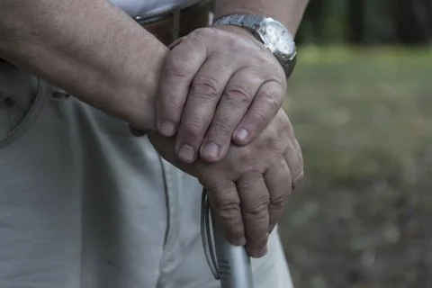 The large hands of an elderly man are folded on a cane with a watch on his ri Stock Photos