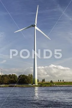 Large metal wind turbine on riverbank with blue sky and clouds Stock Photos