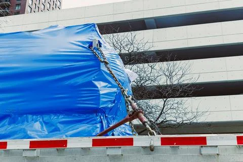 A large object wrapped in blue plastic is secured on a flatbed truck with cha Stock Photos