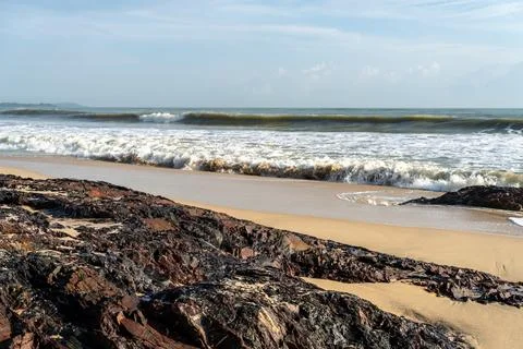 Large ocean waves break and foam onto a sandy beach with dark, jagged rock .. Stock Photos
