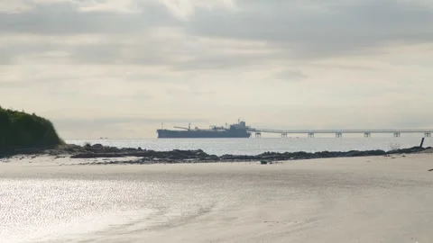 Large ore ship docked at coastal port seen from rocky beach on sunny day Stock Footage 308162957