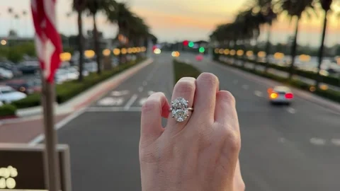 Large oval diamond solitaire engagement ring on hand with cars at sunset Stock Footage 325876532