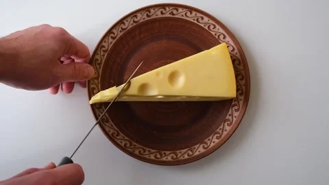 A large piece of cheese lies on a ceramic plate, man cuts a small piece Stock Footage 168528864