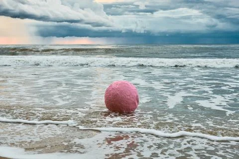 Large pink sphere lapped by waves against seashore against horizon and stor.. Stock Photos
