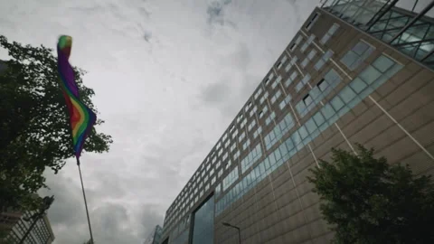 Large rainbow pride flag waves on a tall pole in front of a modern high-rise Stock Footage 346240362