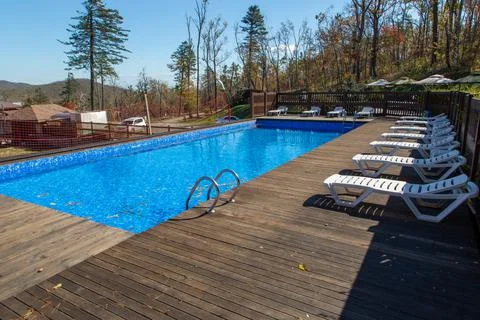 A large rectangular pool with blue water and dry leaves. White lounge chairs lie Foto stock