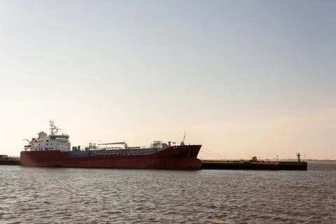 Large red cargo ship floats calmly on water. Sunset hues glow softly behind Stock Photos