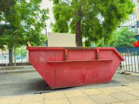 Large red dumpster placed on the street under green trees near buildings du.. Stock Photos