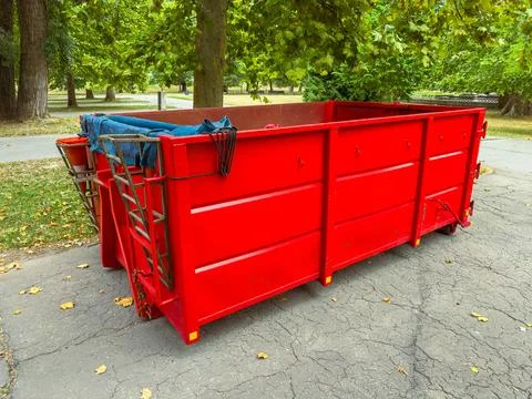 A large red dumpster sits on a paved path in a park where trees surround the  Stock Photos