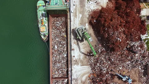 Large ship unloading waste at a recycling facility near a river in a busy urban 스톡 동영상 337902045