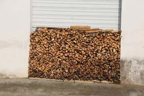 Large stack of split firewood piled against a white wall beneath a roll-up do Stock Photos