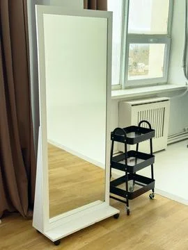 Large standing mirror with a simple design near a window and a small cart i.. Stock Photos