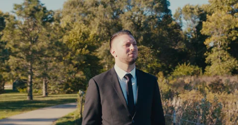 A large, strong-built man standing in a jacket and white shirt against a nature 스톡 동영상 321621371