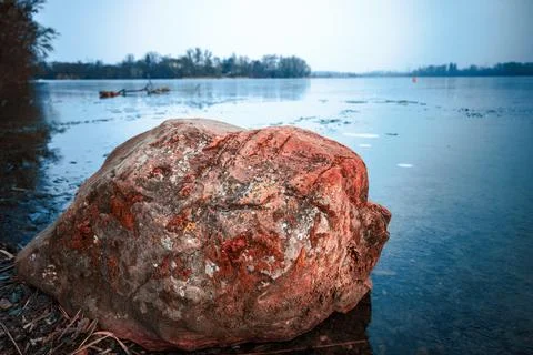 A large, textured red boulder rests on the shoreline of a partially frozen la Stock Photos
