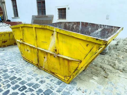 A large yellow dumpster sits on a cobblestone street next to a construction s Stock Photos