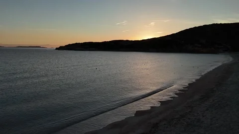 Lateral Drone Sweep Over Calm Beach with Sun Peeking Behind Coastal Hill Vídeo Stock 334796968