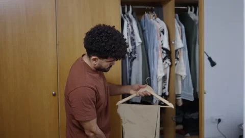Latin man opens his wardrobe and takes out a pair of camel trousers and a white Stock-Footage 257595128