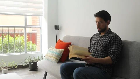Latin man sitting on a couch leafing a text book in a concentrated attitude Stock Footage 340583357