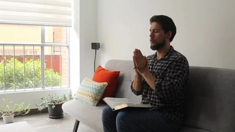A latin man sitting on a couch praying with a rosary and a holy bible Vídeo Stock 340583485