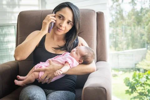 Latin mother using talking on cell phone with her sleeping baby in her arms Stock Photos
