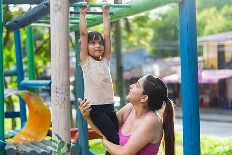 Latina mother smiling while supporting her daughter as she hangs from monke.. 写真素材