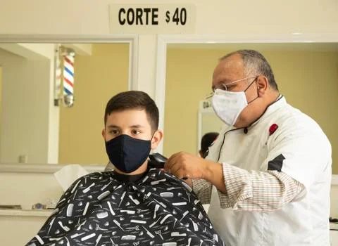 Latino barber cutting hair of teenage boy, wearing protection mask due to pan Stock Photos