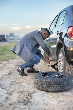 Latino man on suit changes a flat tire on his car at the side of the road. Stock Photos