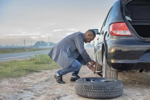 Latino man on suit changes a flat tire on his car at the side of the road w.. Stock Photos