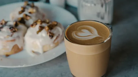 Latte leaf design in glass cup next to fresh icing buns serving cappuccino Stock Footage 132569919