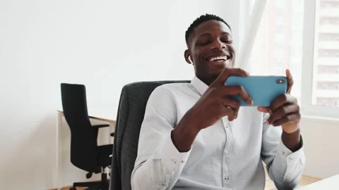 A laughing black guy with earbuds is looking to his smartphone in the office Stock Footage 154361004