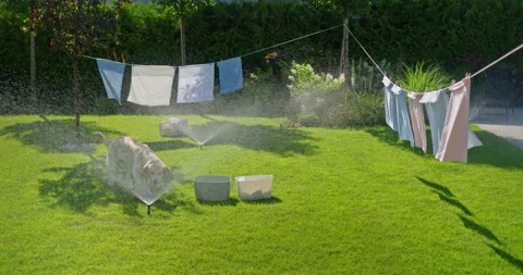 Laundry drying on a clothesline gets soaked by automatic sprinklers over a green Stock Footage 281556628