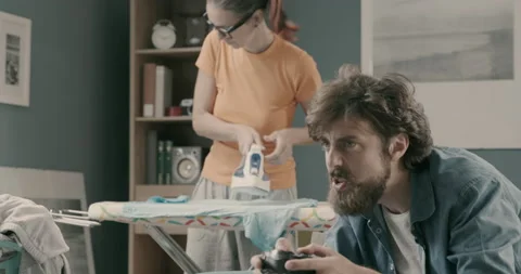 Lazy husband playing videogames while his wife is ironing Stock Footage 151100613