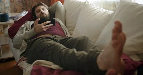 Lazy man beard lying on the couch napping, watching bored content on smartphone Stock Footage 238418414