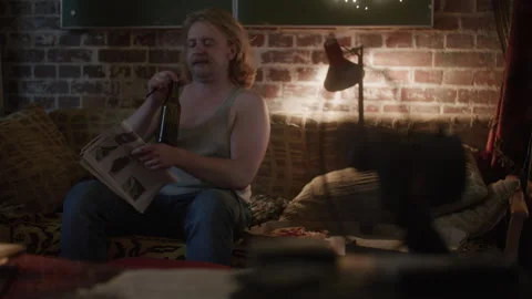 A lazy man sitting on a couch in his apartment and drinking beer while read.. Video stock 259688218