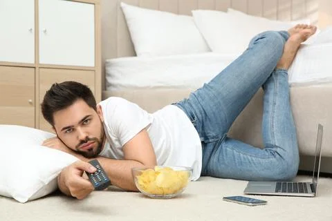 Lazy young man with bowl of chips watching TV while lying on floor at home 写真素材