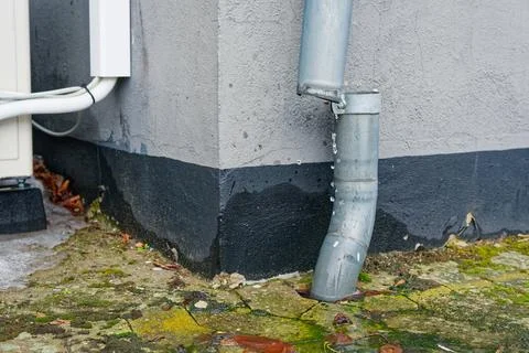 Leaking metal downspout pipe draining water onto mossy concrete ground near.. Stock Photos