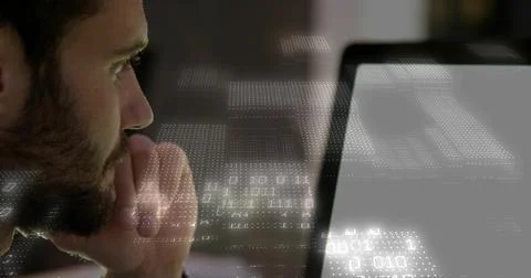 Leaning bearded man studying bright laptop at desk in dark sweater with binary 스톡 사진