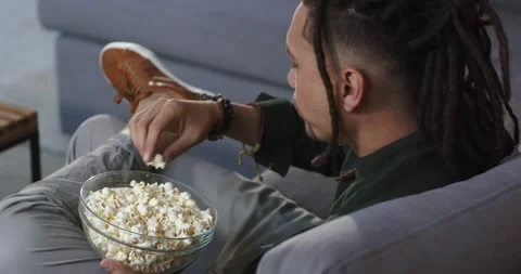 Leaning forward to snack, man is dipping hand into bowl and eating popcorn on Stock Footage 336784141