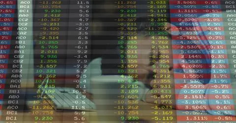 Leaning man studying financial data overlay on laptop at desk, wearing red blue Foto stock