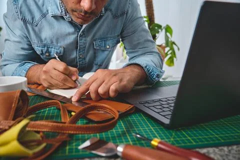 Leather craftsman taking notes and planning work using laptop and notebook Stock Photos