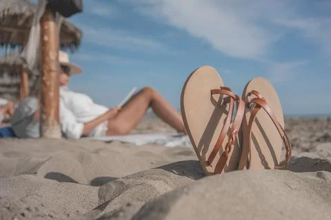 Leather sandals standing on the beach while tourist relaxes reading a book Stock Photos