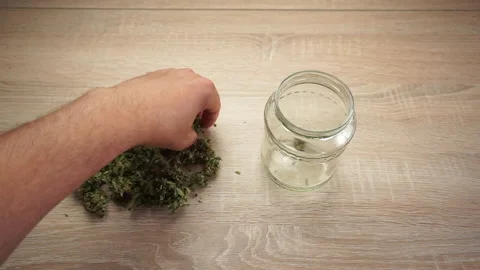 Left hand filling glass jar with marijuana buds on wooden table Stock Footage
