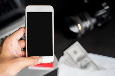 Left hand holding smartphone on blur blackground with laptop, dollars, camera Stock Photos