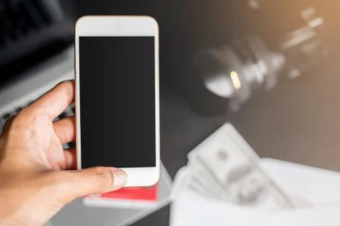 Left hand holding smartphone on blur blackground with laptop, dollars, camera Stock Photos