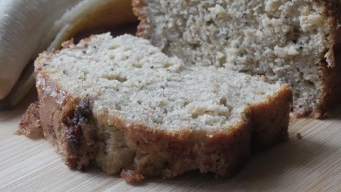 Left pan view of banana bread cut in half on a wooden board with a bananas in 스톡 동영상 296574674